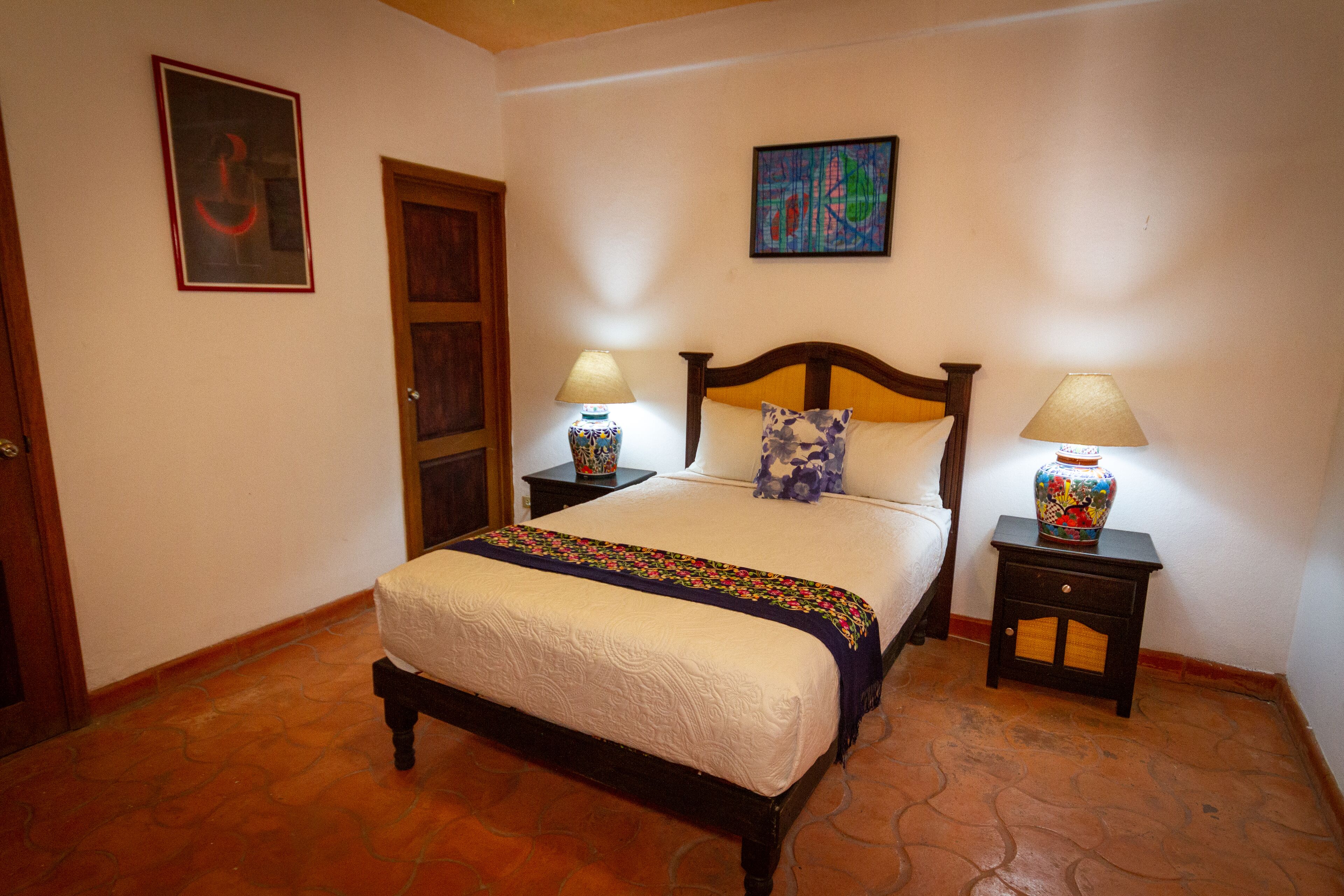 A bedroom with a large bed, two bedside tables with lamps, a door, and two wall-mounted paintings.