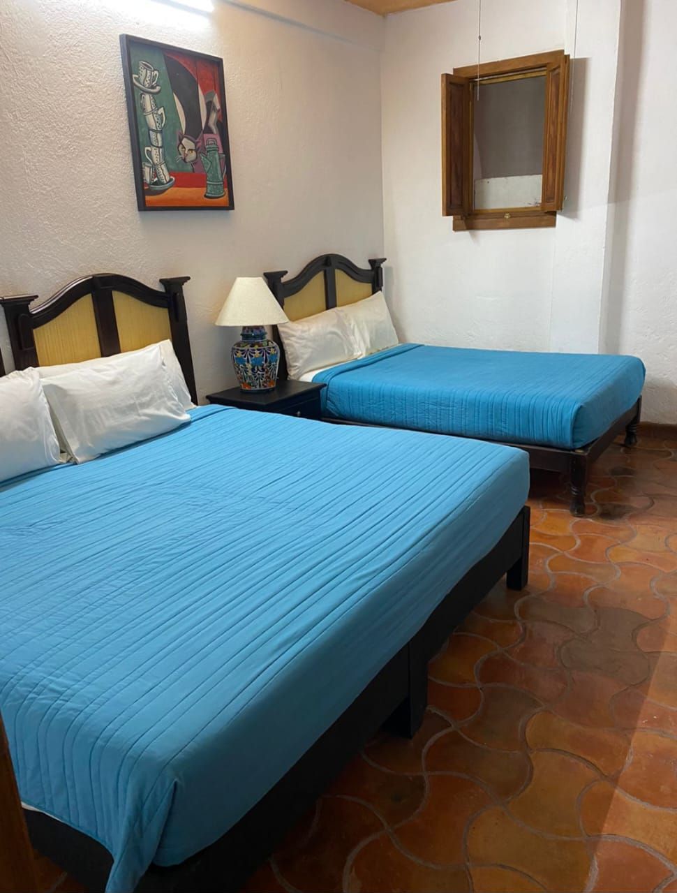 Una habitación con dos camas, cada una con ropa de cama azul, una mesita de noche con una lámpara, un cuadro en la pared y un espejo sobre la cama.