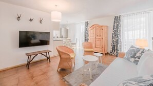 Ein modernes Wohnzimmer mit Flachbildfernseher, Rattanmöbeln, einem Holzkabinett und einem großen Fenster mit gemusterten Vorhängen.