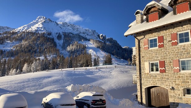 Ein Steinhaus mit verschneietem Dach und Holzläden neben einer verschneiten Straße mit parkenden Autos.