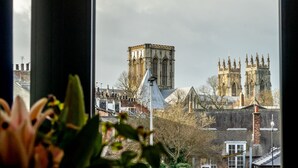 Vista de un edificio histórico con arquitectura gótica, que incluye arcos apuntados y una aguja, visible a través de una ventana con una flor en primer plano.