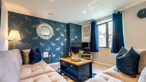 Una sala de estar moderna con pared azul con estampado de ciervo, un espejo redondo, un televisor y una mesa de centro de madera.