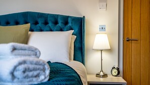 Una cama bien hecha con un cabecero verde azulado, una lámpara de noche y un reloj sobre una mesita de noche de madera.