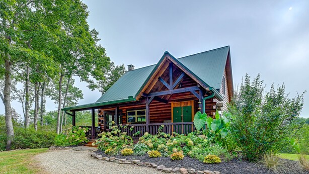 A log cabin with a green roof, surrounded by a gravel driveway and a garden with various plants. - Mtn Views, Yard, & Fire Pit: Multi-Level Cabin (Franklin)