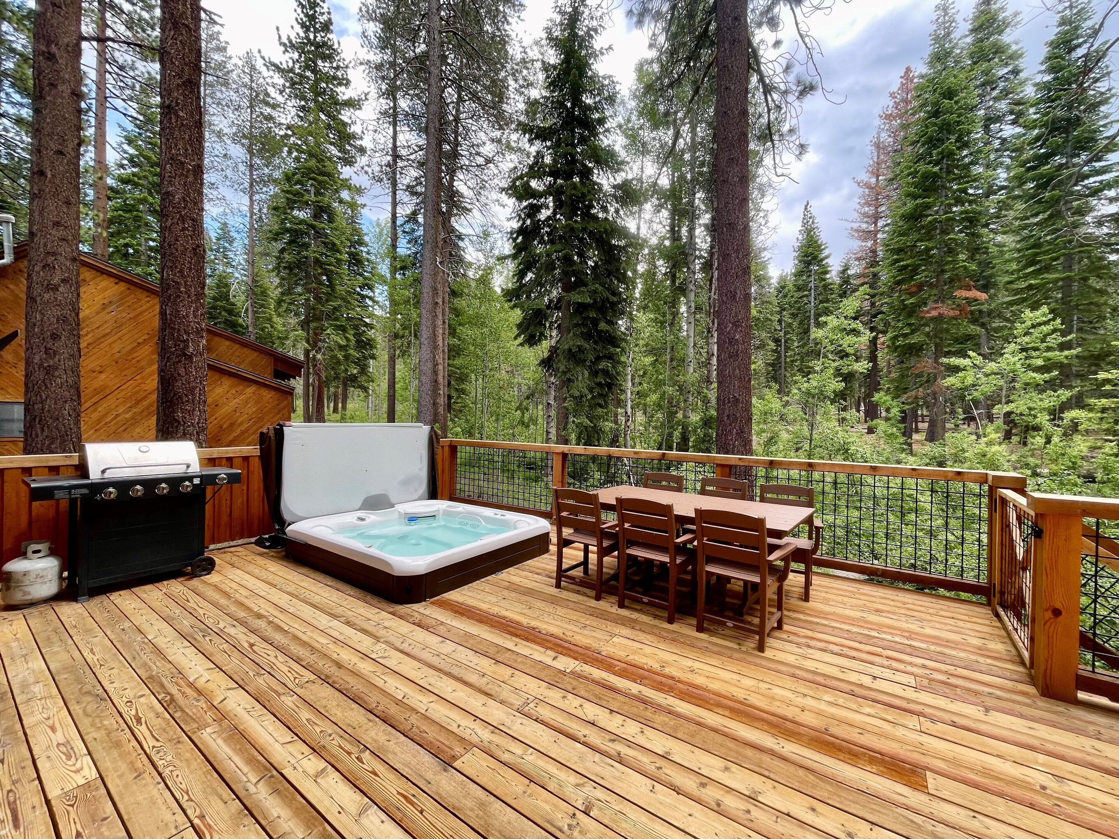 A wooden deck with a hot tub, a grill, and a table with chairs, surrounded by tall trees.