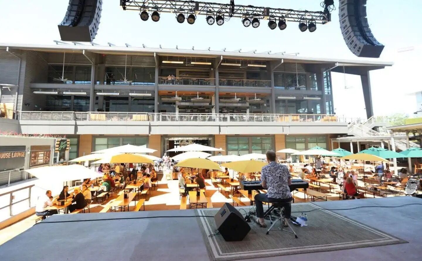 Un espace de restauration en plein air avec plusieurs tables et des parasols, une scène équipée de matériel de sonorisation et, au fond, un bâtiment moderne doté de balcons.