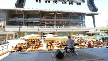 Un espace de restauration en plein air avec plusieurs tables et des parasols, une scène équipée de matériel de sonorisation et, au fond, un bâtiment moderne doté de balcons.