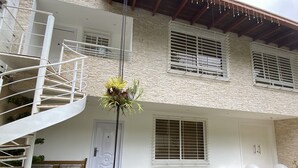 Un edificio de dos pisos con fachada blanca, una escalera espiral y un pequeño jardín con un banco y plantas en macetas.