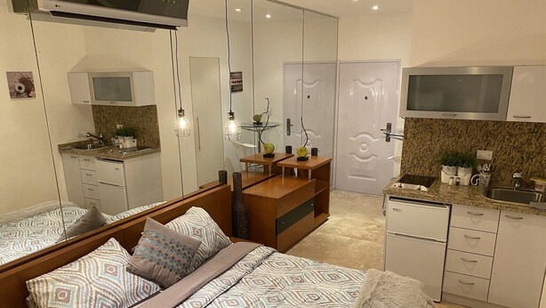 Un dormitorio moderno con cama, mesitas de noche, espejo y una kitchenette con microondas y fregadero.