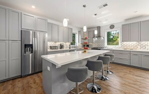 A modern kitchen with grey cabinets, a stainless steel refrigerator, a large island with bar stools, and pendant lighting. - Miramar Beach House. A short walk to the beach. (Half Moon Bay)