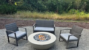 Een ronde vuurkuil met stenen en een label, omringd door drie rieten stoelen met kussens.