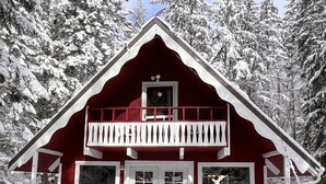 Una cabaña roja con balcón, detalles blancos y un bosque nevado de fondo.