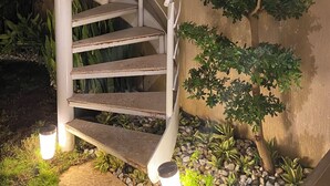 Un escalier en colimaçon avec une rampe blanche, entouré d’un jardin où poussent différentes plantes et agrémenté d’un arbre fixé au mur.