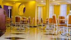 Un comedor de hotel con sillas de metal blanco, paredes amarillas y una decoración circular grande en la pared.