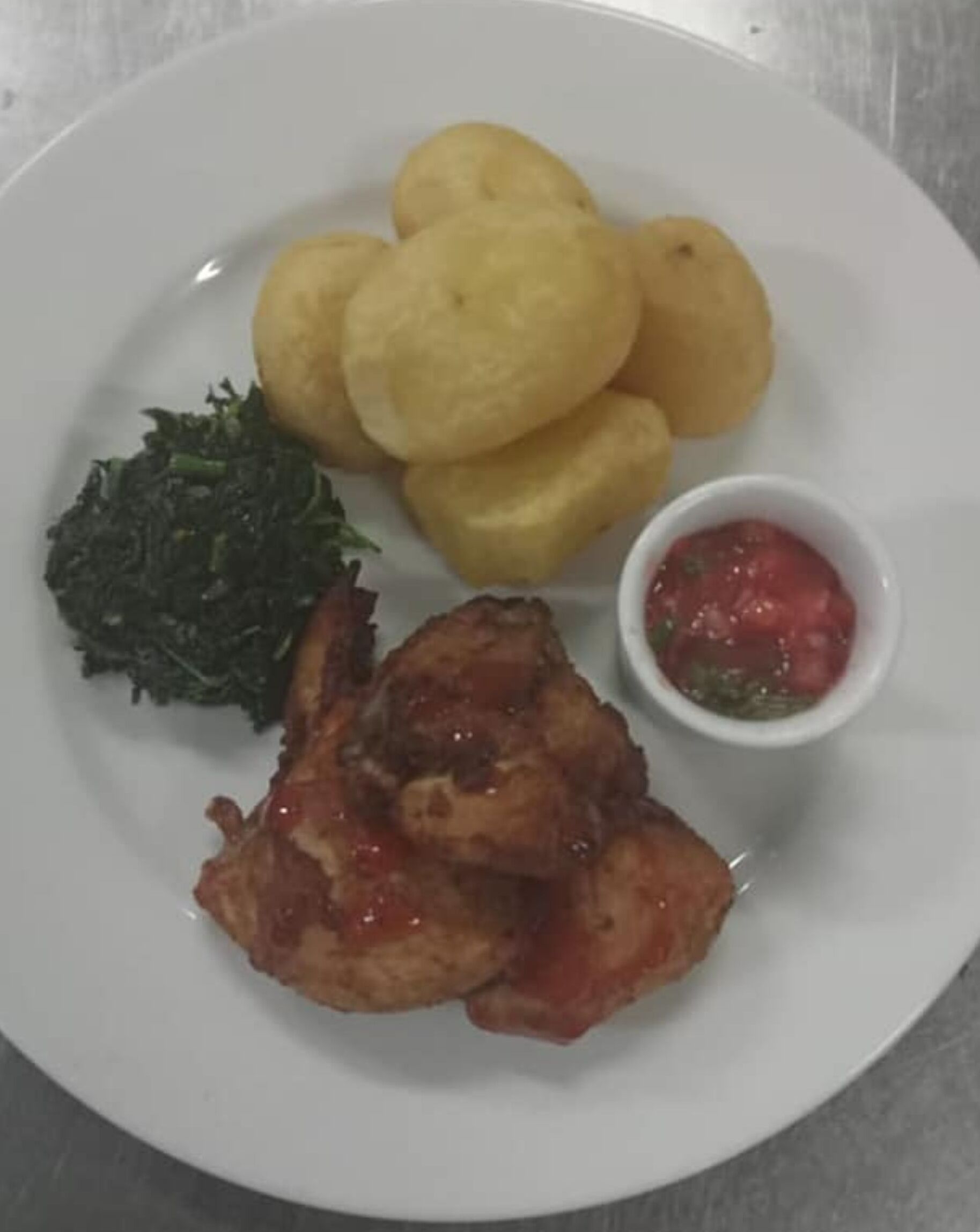 A plate with fried chicken, fried plantains, greens, and a small bowl of sauce.