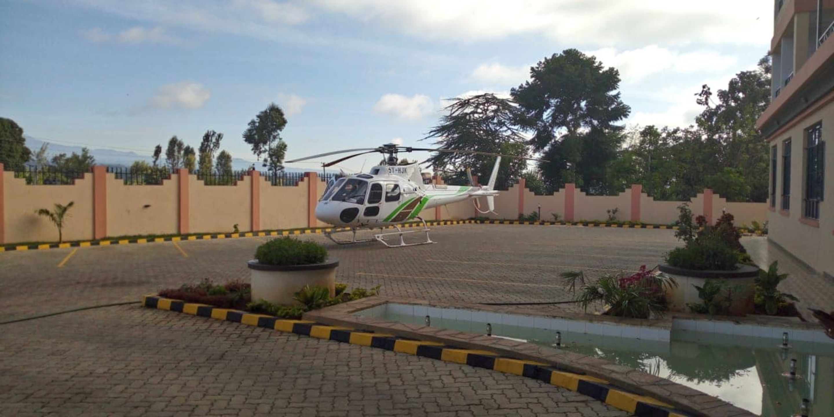 Un helicóptero estacionado en un patio con una zona pavimentada, una piscina pequeña y un edificio al fondo.