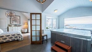 A bedroom with a large bed, a wooden door, a window with a view of the sea, and a hot tub.