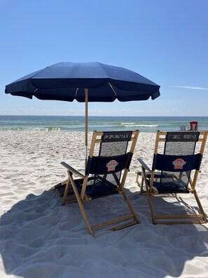 Zwei Strandstühle mit einem blauen Sonnenschirm am sandigen Strand.