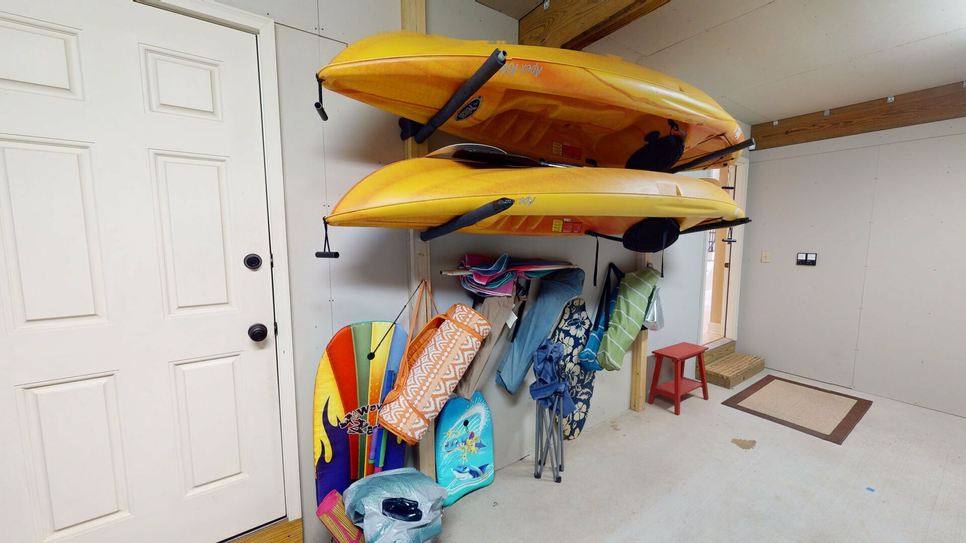 A room with two kayaks mounted on the wall, a colorful surfboard, and a small red stool.