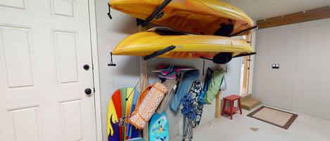 A room with two kayaks mounted on the wall, a colorful surfboard, and a small red stool.