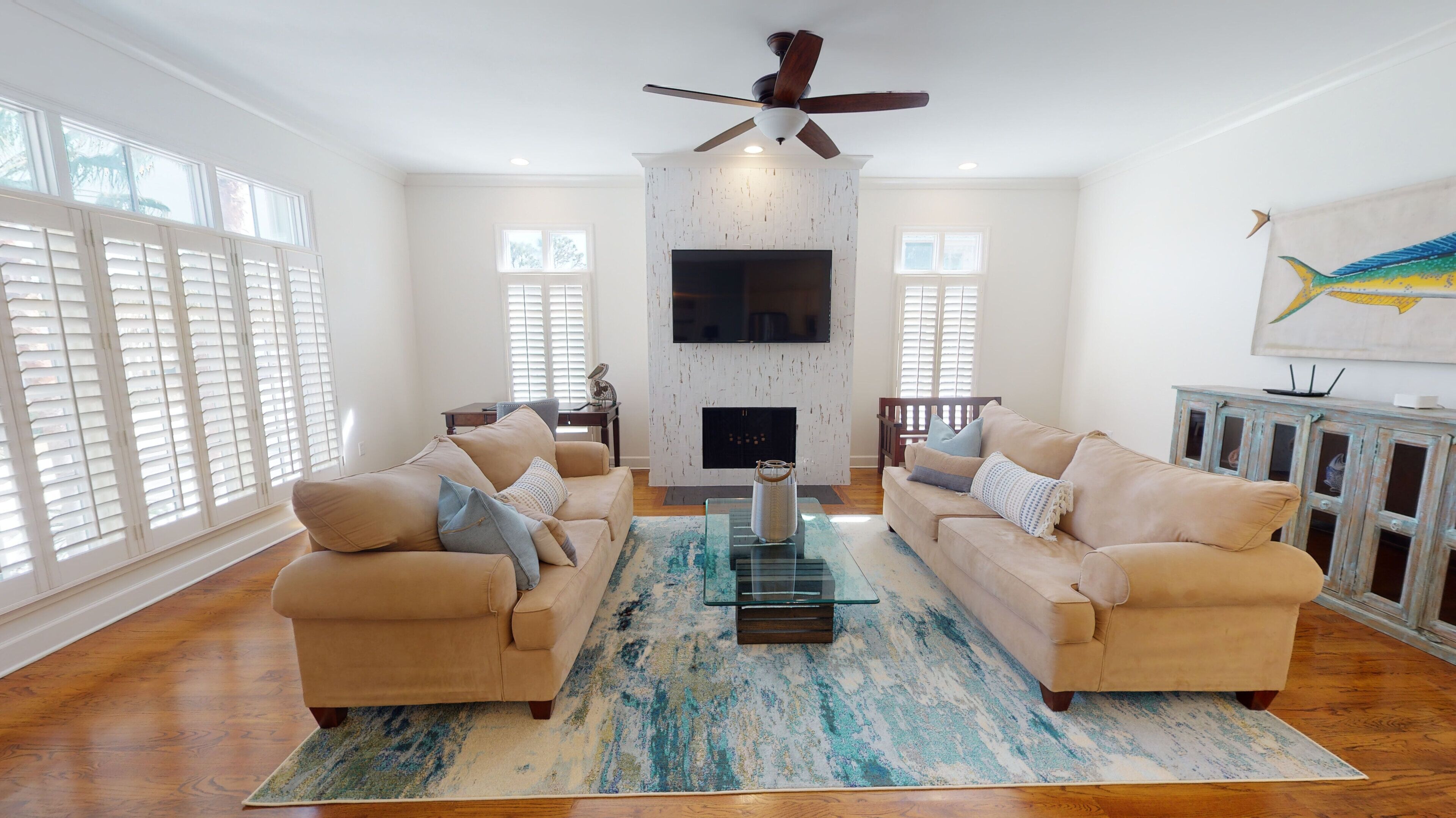 A living room with two beige sofas, a glass coffee table, a fireplace, and a ceiling fan.