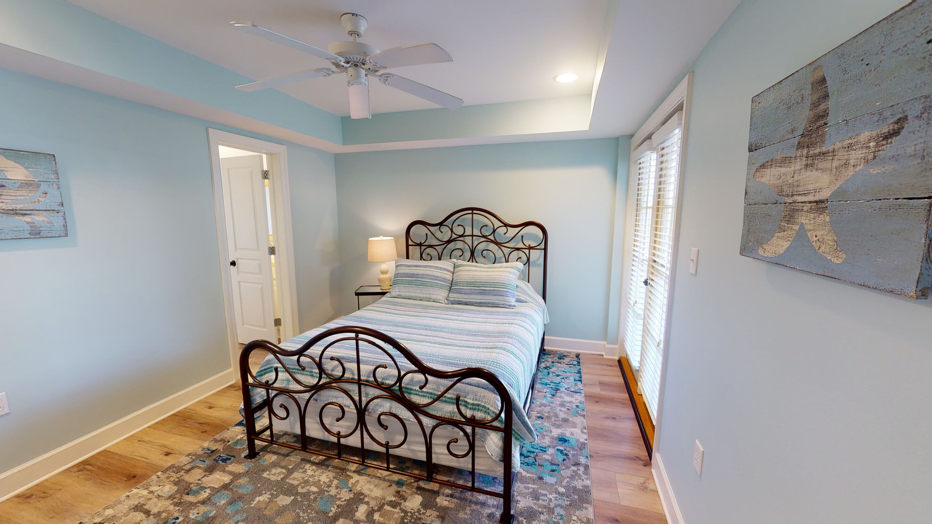 A bedroom with a bed, a ceiling fan, a door, a nightstand, a lamp, and a wall with a decorative piece.