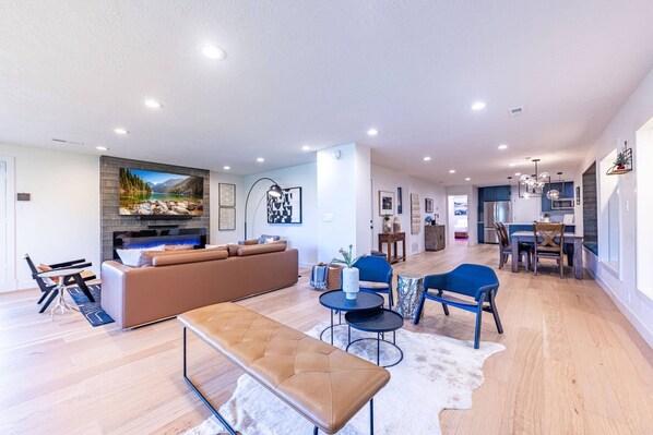A spacious living area with a large sofa, a coffee table, and a bench. There is a dining area with a table and chairs, and a kitchen in the background.