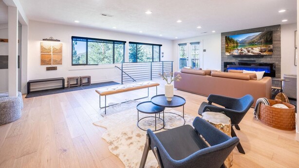Een ruime woonkamer met een grote bank, twee fauteuils, een salontafel en een open haard. Grote ramen bieden uitzicht op bomen en een meer.