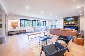 A spacious living area with a large sofa, two armchairs, a coffee table, and a fireplace. There are large windows offering a view of trees and a lake.