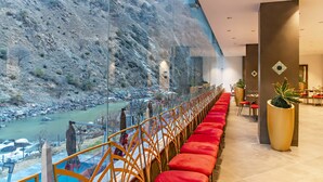 A hotel lobby with a long red bench, a large potted plant, and a view of a river through a glass wall. - Echor Palm Bliss Riverside Resort & Spa Kasol (Bhuntar)