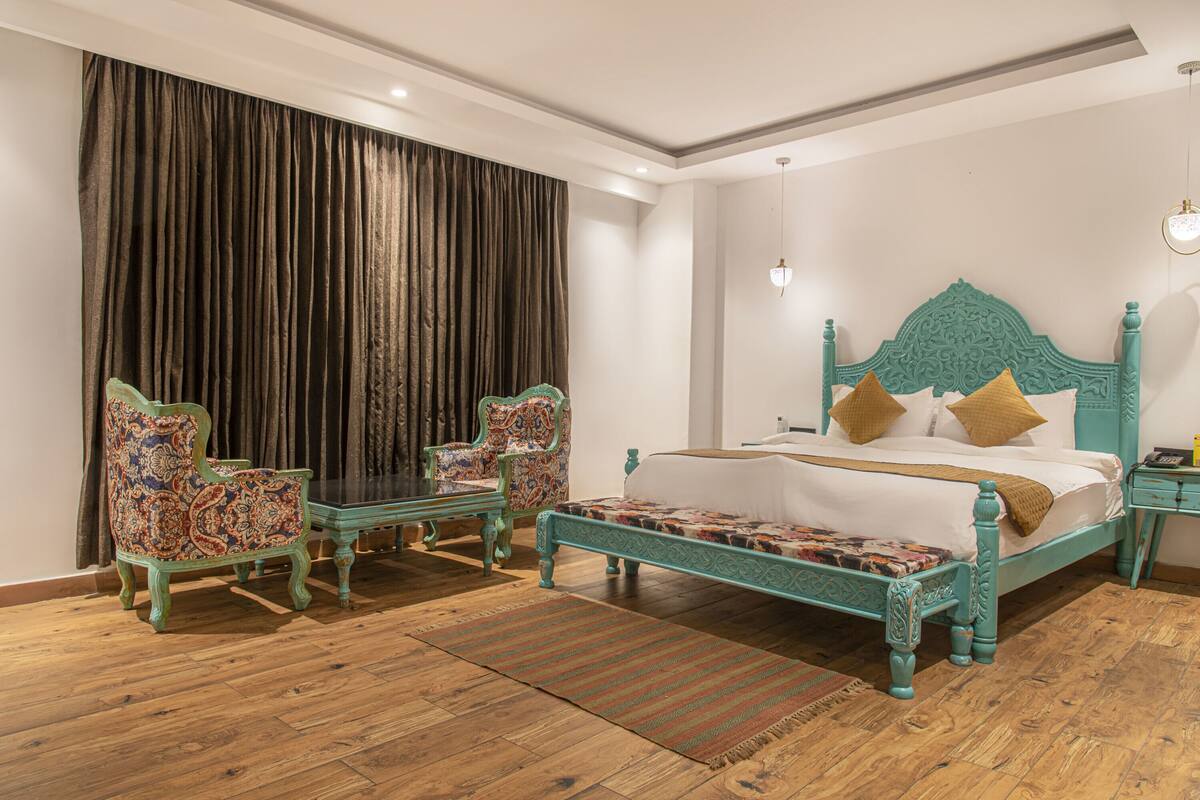 Quarto com cama, duas cadeiras, mesa de cabeceira e uma janela grande com cortinas.