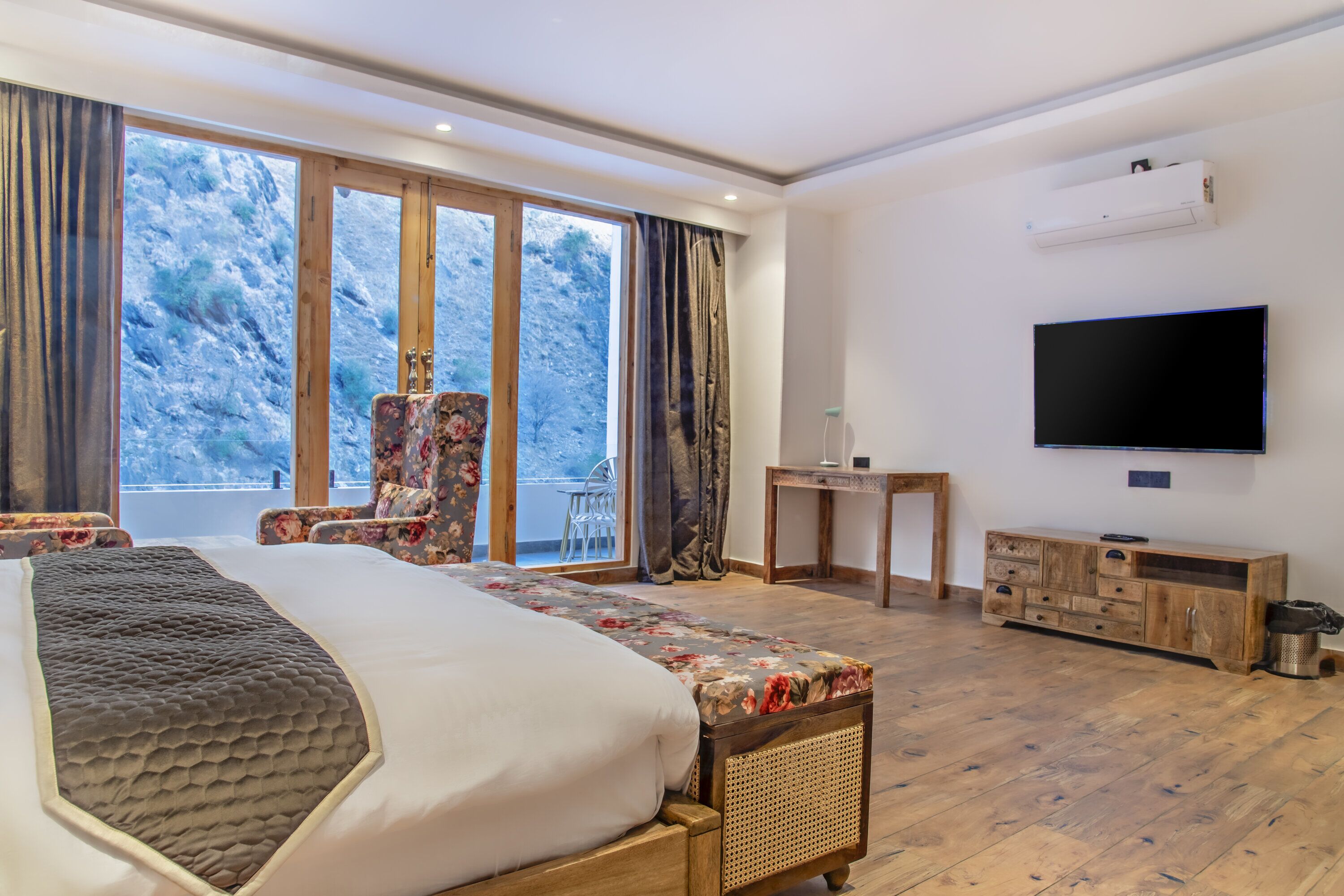 A spacious bedroom with a large bed, a TV, a desk, and a view of a mountainous landscape through the windows.