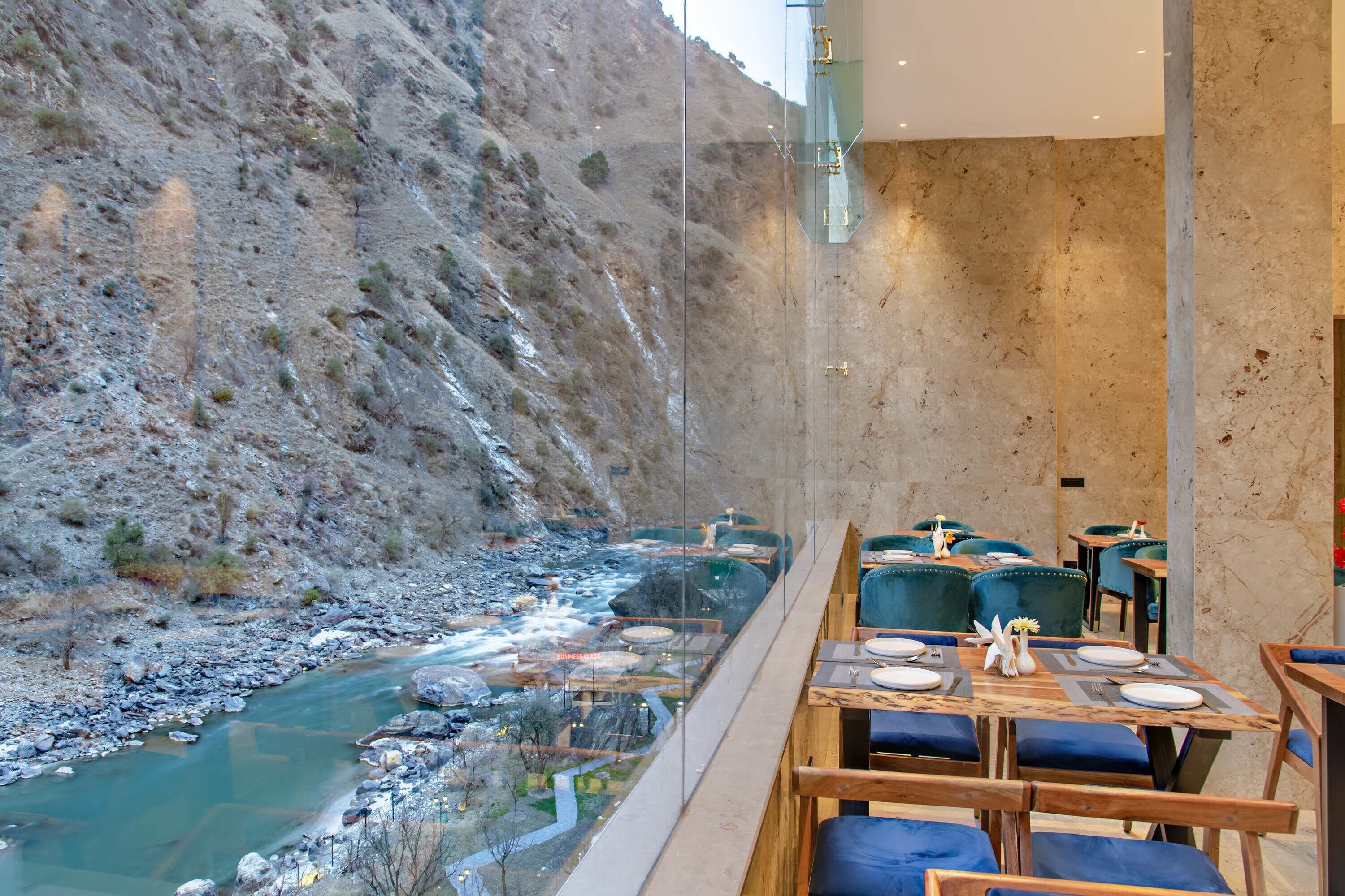 Un restaurante con vista a un río y montañas, mesas de madera y sillas azules, y un diseño interior moderno.