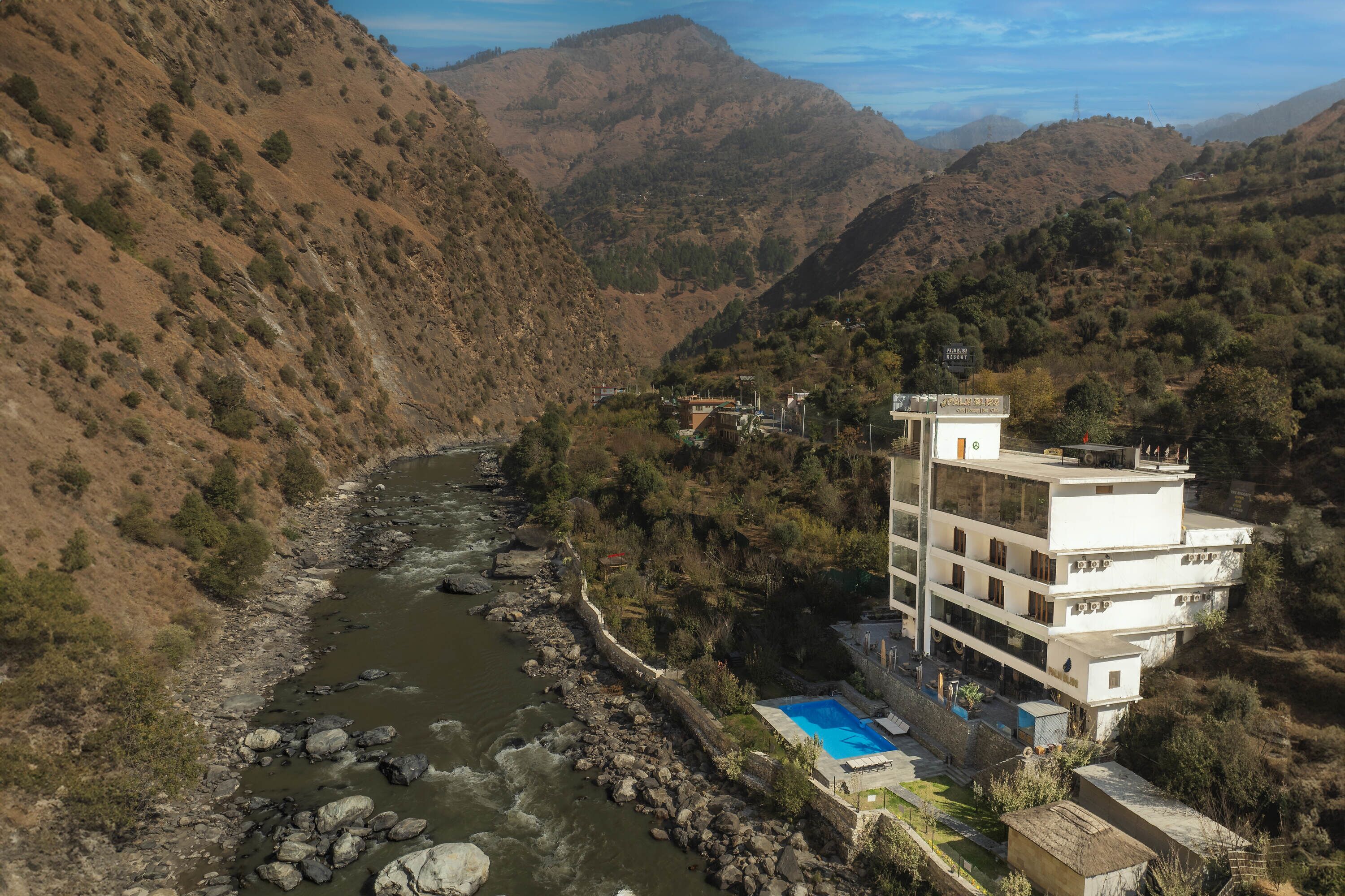 Un hôtel à plusieurs étages doté d’une piscine, situé dans une vallée, avec une rivière et des montagnes en arrière-plan.