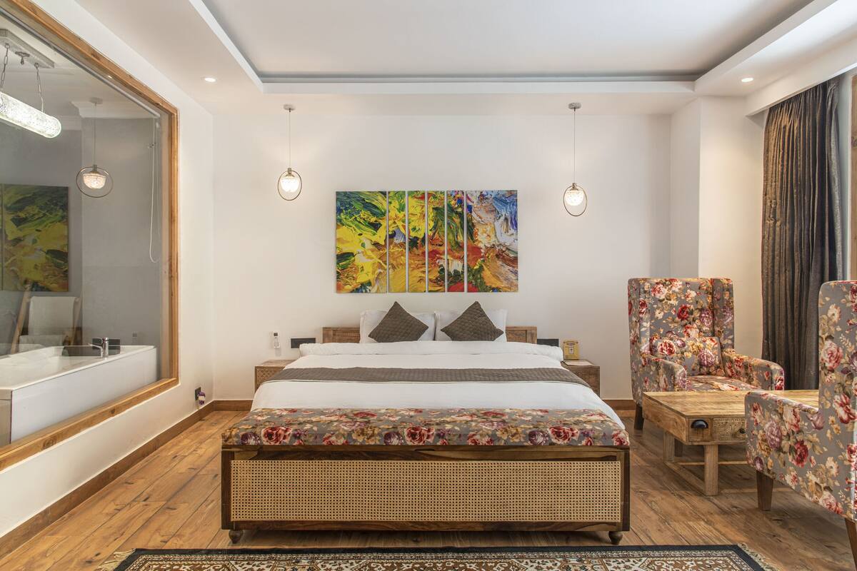 Quarto com uma cama grande, um banco de madeira, uma cadeira, um espelho grande e uma pintura na parede.