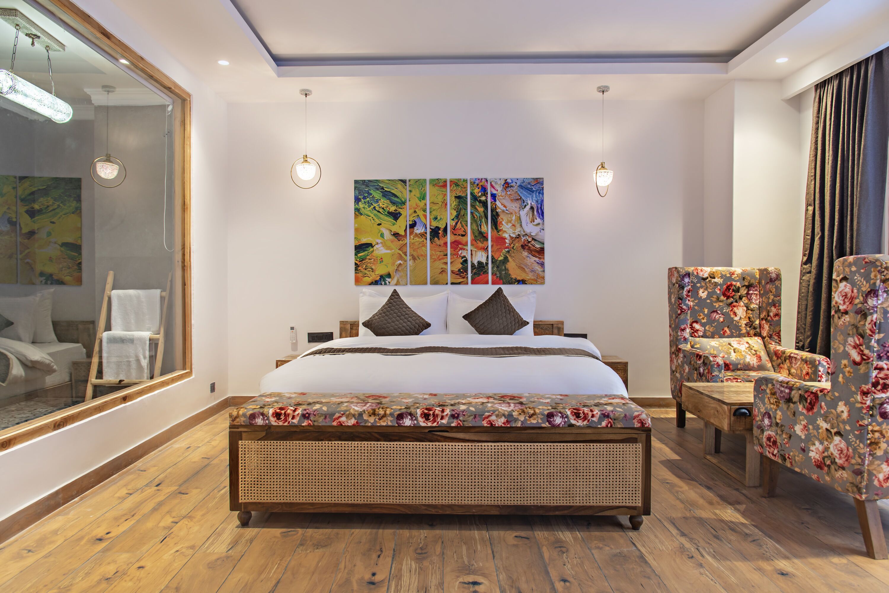 A modern bedroom with a large bed, a wooden floor, a large mirror, a painting on the wall, and a floral armchair.