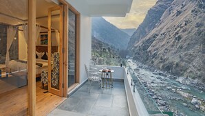 A balcony with a view of a river and mountains, a dining area with a table and chairs, and a bedroom with a bed and curtains. - Echor Palm Bliss Riverside Resort & Spa Kasol (Bhuntar)