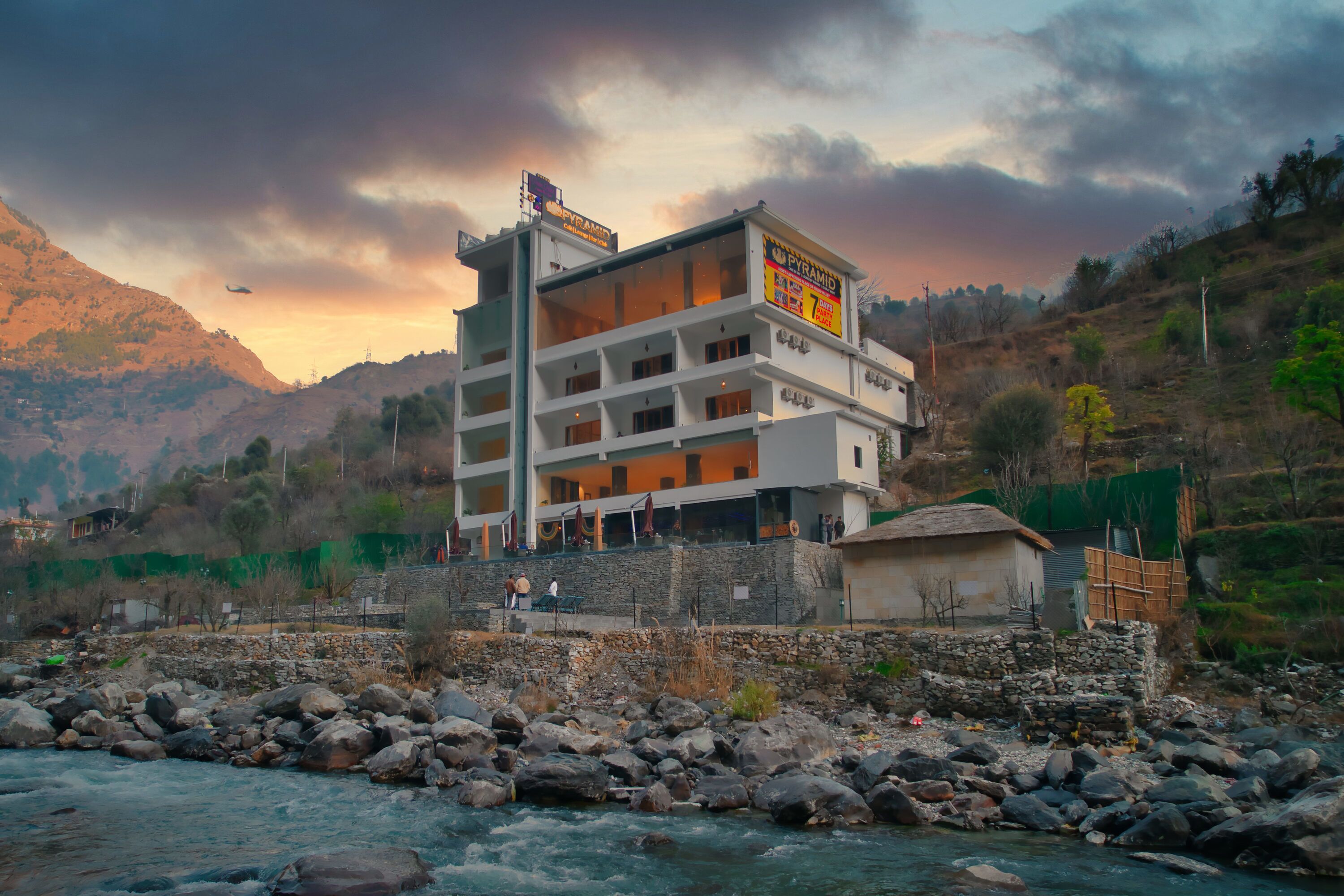 A multi-story hotel with balconies, located near a river with rocky terrain and mountains in the background.