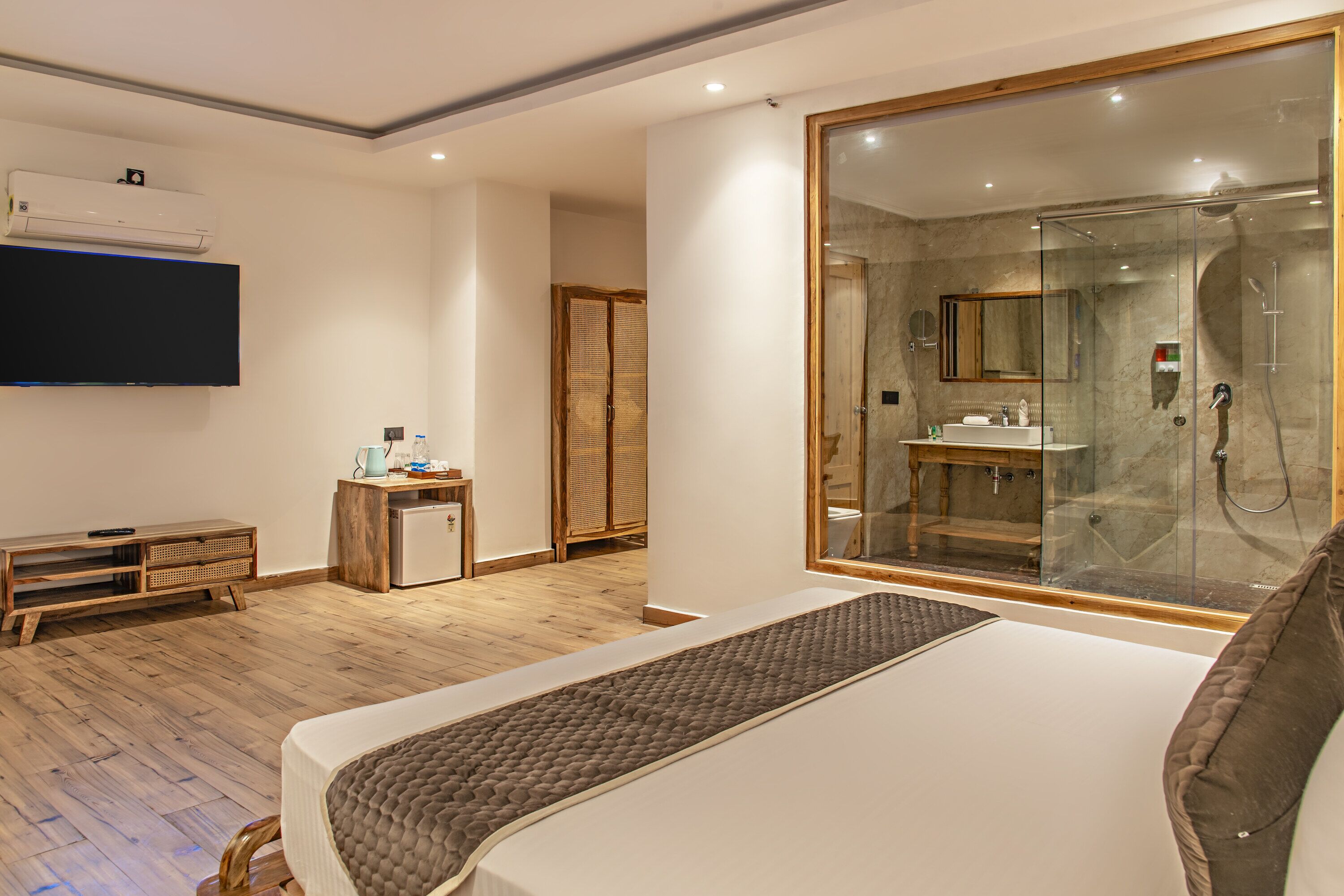 A modern hotel room with a large bed, a glass-enclosed shower, a flat-screen TV, and a wooden floor.