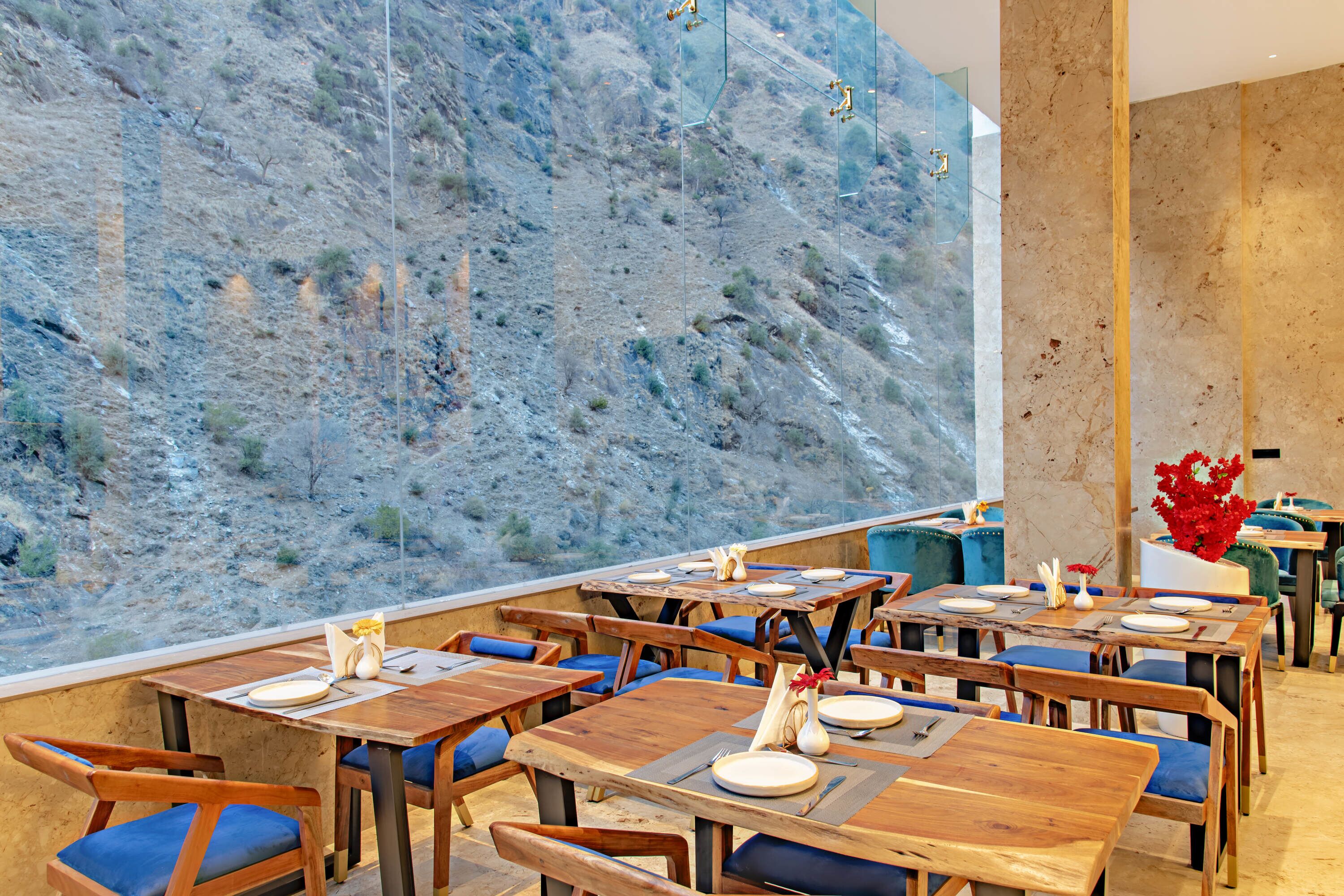 A restaurant with wooden tables and blue cushioned chairs, a large rock wall, and a vase with red flowers.