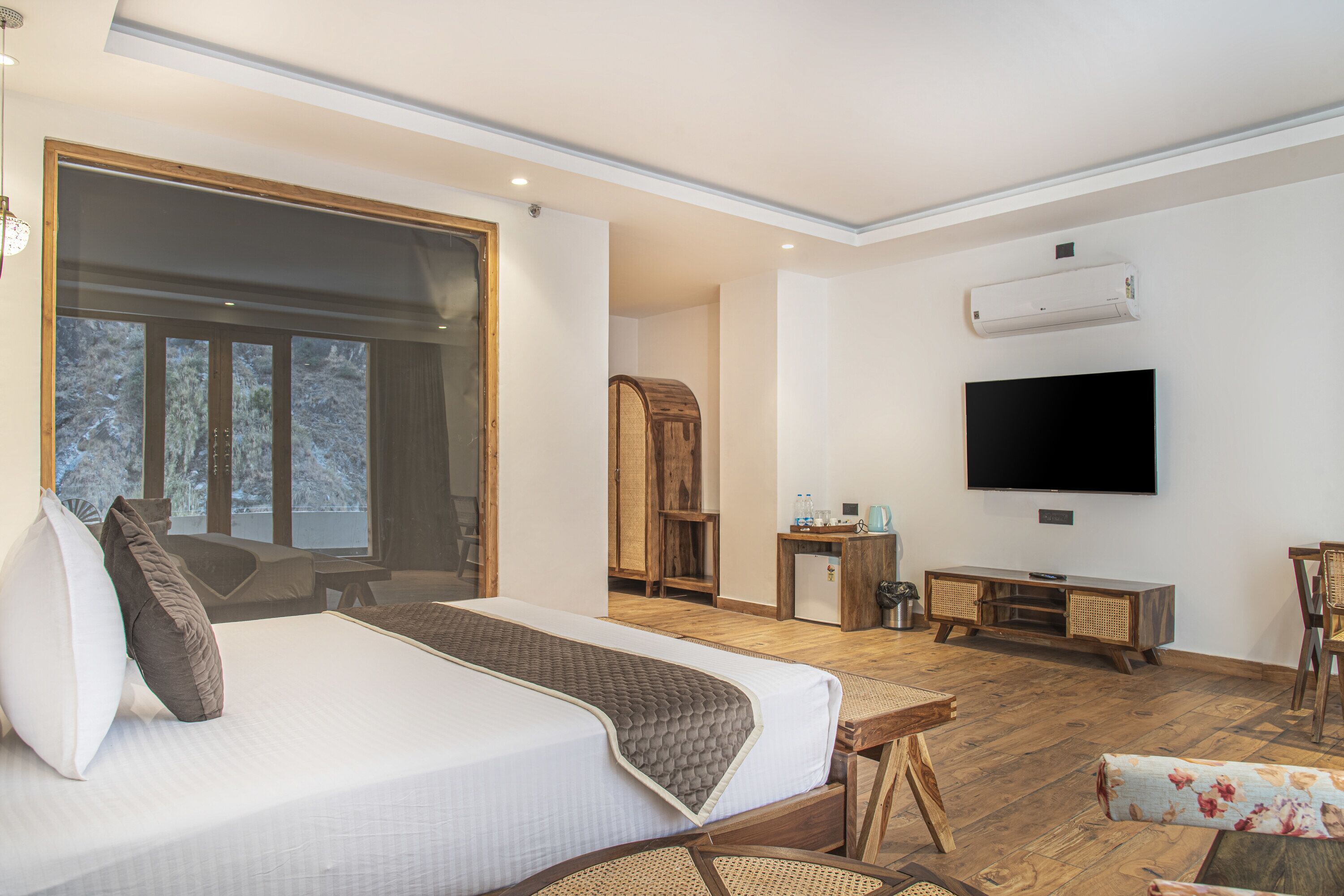 A modern hotel room with a large bed, a flat-screen TV, a wooden desk, and a view of trees through the window.