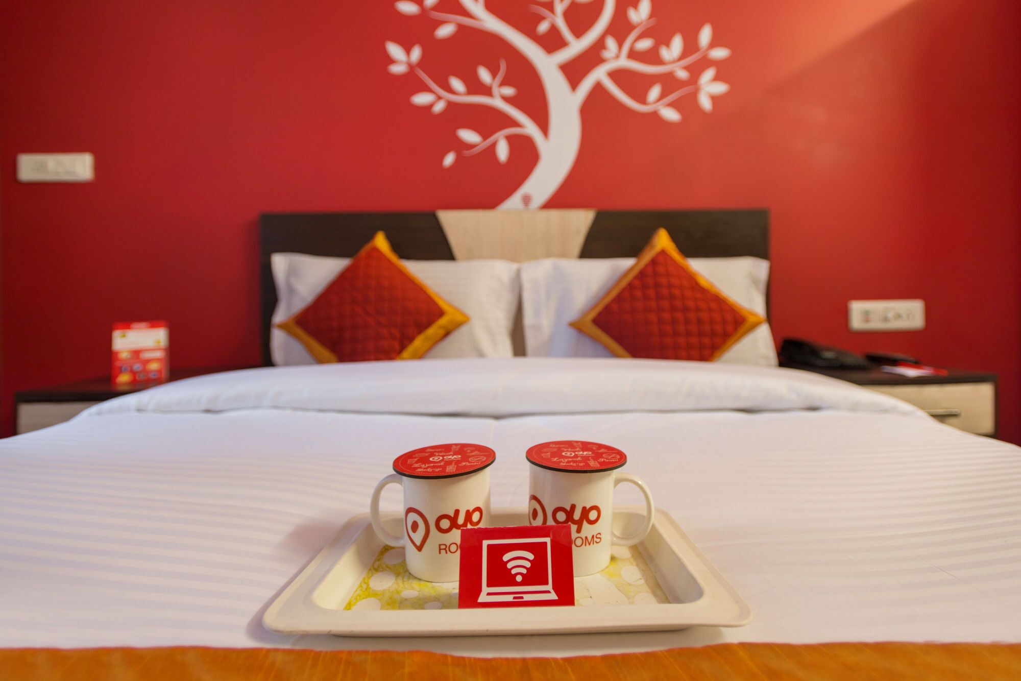 A hotel room with a bed, two pillows, a tray with two coffee mugs, and a wall decoration.