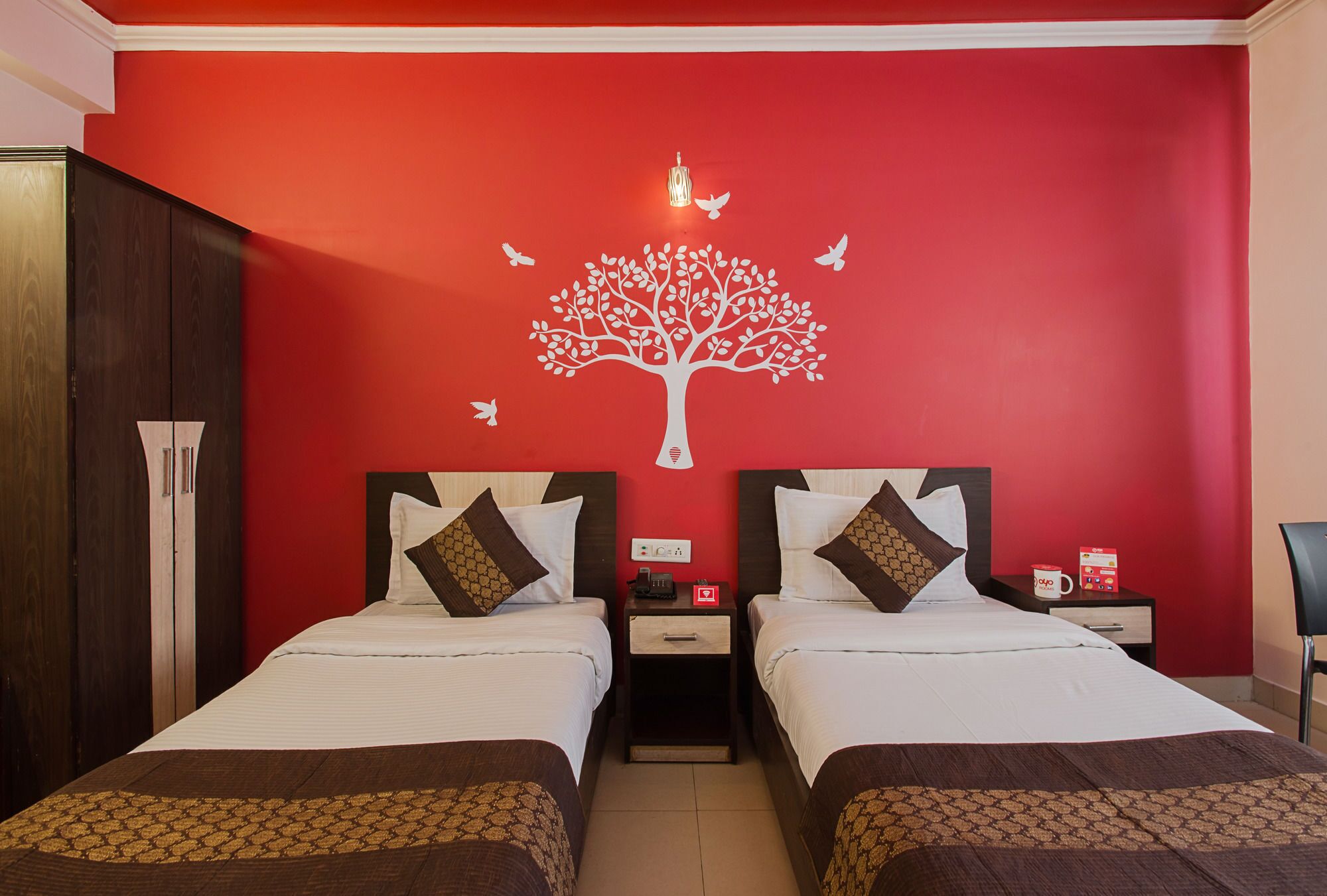 Habitación de hotel con dos camas, una pared roja con un mural de un árbol blanco y una mesita de noche con un teléfono y una caja roja.