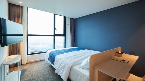 Quarto de hotel com cama, escrivaninha, televisão na parede e uma grande janela com vista.