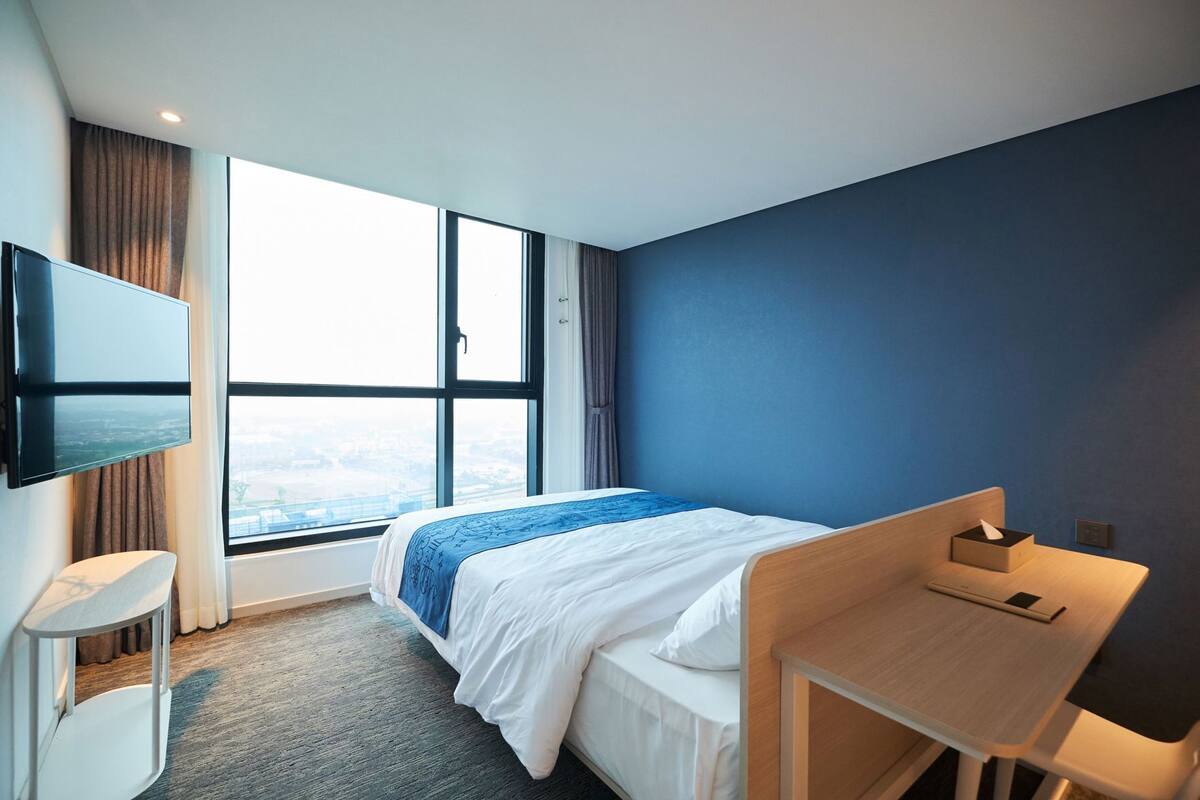 Une chambre d’hôtel équipée d’un lit, d’un bureau, d’un téléviseur fixé au mur et d’une grande fenêtre donnant sur la ville.