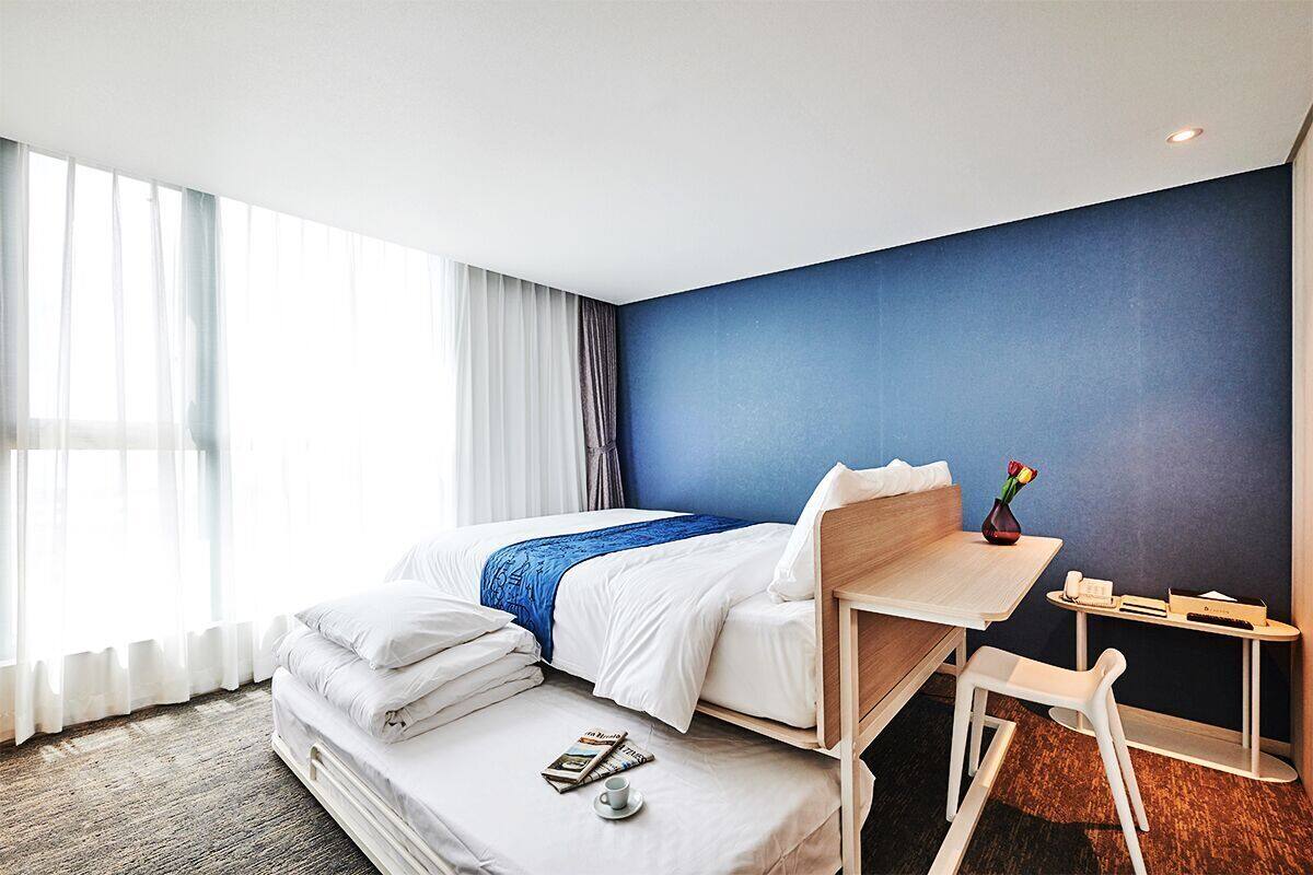 Une chambre d’hôtel avec un lit, un bureau, une chaise, une petite table et une fenêtre avec des rideaux.