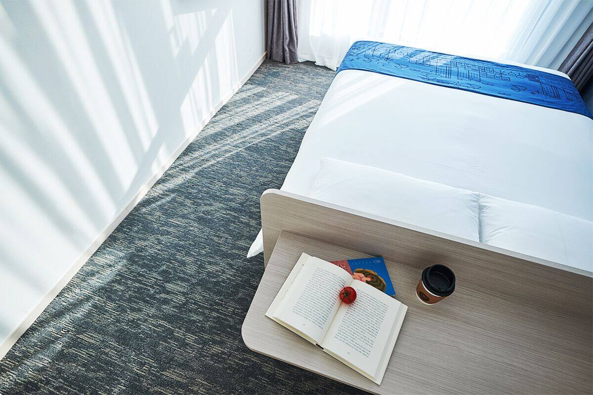 Un lit bien fait, avec une tête de lit bleue, une table d’appoint sur laquelle reposent un livre et une tasse de café, et une fenêtre avec des rideaux.
