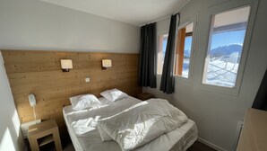 Ein Schlafzimmer mit einem hölzernen Kopfende, zwei Nachttischlampen, einem Bett mit weißer Bettwäsche, einem Nachttisch, einem Holzschemel und einem Fenster mit Vorhängen.