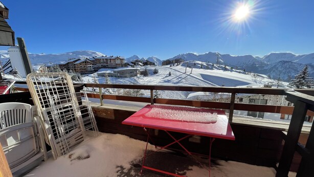 Ein Balkon mit einem roten Tisch und Metallstühlen, mit Blick auf eine verschneite Berglandschaft mit Gebäuden und Skipisten.