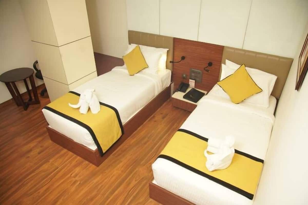 A hotel room with two beds, each with a wooden headboard, white bedding, and yellow accents. There is a small table and a chair in the corner.